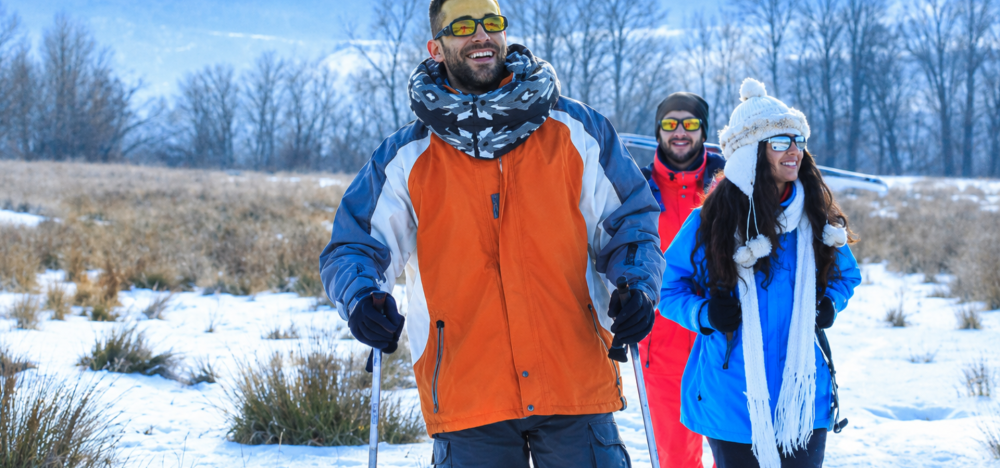 Three people snowshoeing in winter landscape, wearing colorful jackets and sunglasses.
