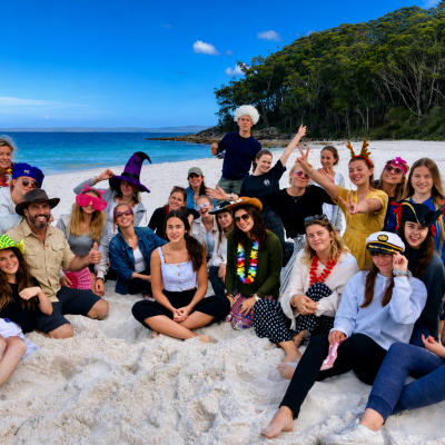 Group of people in costumes posing on a beach with trees and ocean in the background.