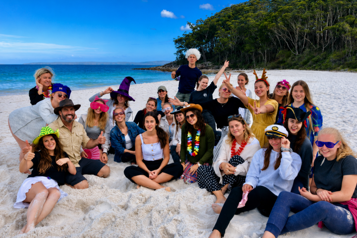 Group of people in costumes posing on a beach with trees and ocean in the background.