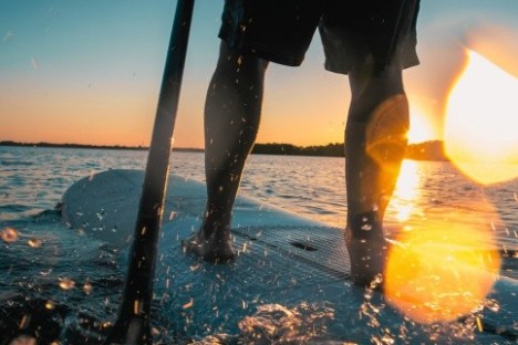 Person paddleboarding at sunset, water splashing, warm glow.