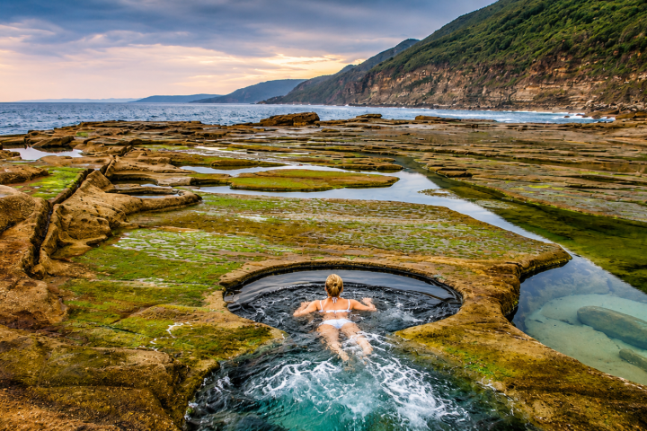 Person in natural rock pool by the ocean, surrounded by rocky terrain and greenery.