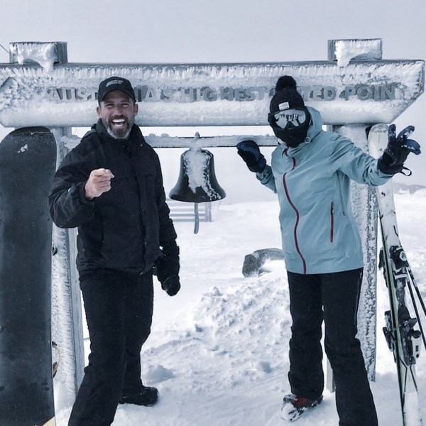 Two people in winter gear at a snowy mountaintop with skis and snowboard, standing by a frosty bell.