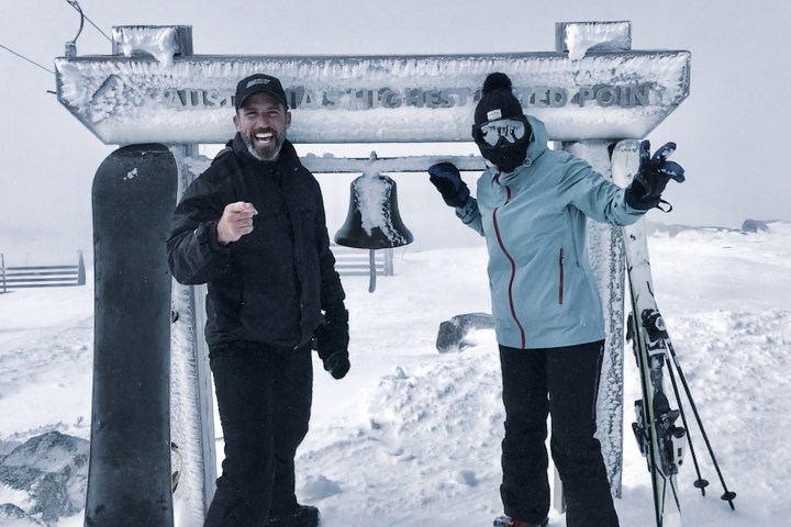 Two people in winter gear at a snowy mountaintop with skis and snowboard, standing by a frosty bell.