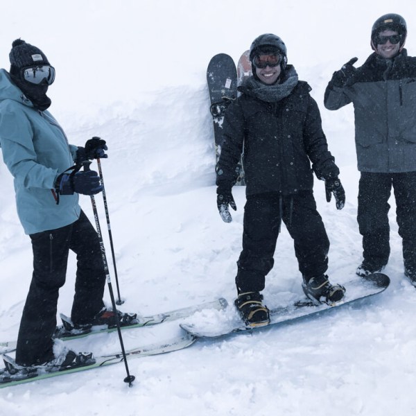Three people in winter gear with skis and snowboards in a snowy landscape.