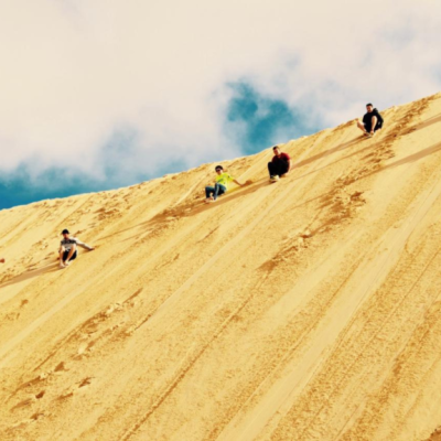 People sliding down a steep sand dune under a blue sky.