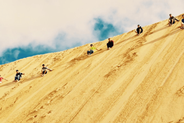 People sliding down a steep sand dune under a blue sky.