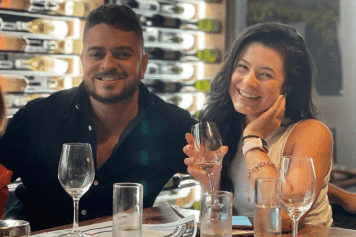 Two people smiling at a table with wine glasses and a wine rack in the background.