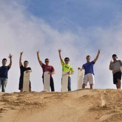 Seven people holding sandboards stand on a sand dune under a blue sky.