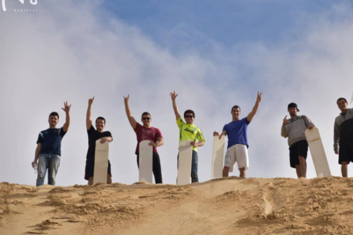 Seven people holding sandboards stand on a sand dune under a blue sky.