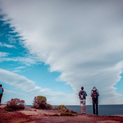 People with backpacks stand on a cliff under dramatic clouds by the sea.