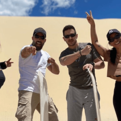 Four people posing with sandboards on sunny dune.
