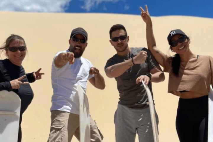 Four people posing with sandboards on sunny dune.