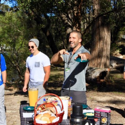 Group of four men having a picnic outdoors, with food and drinks on the table.