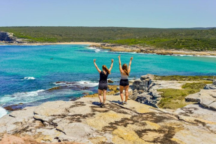 Two people celebrating on a rocky cliff above a turquoise ocean.
