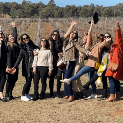 Group of joyful people posing outdoors in front of vineyard, waving and smiling.