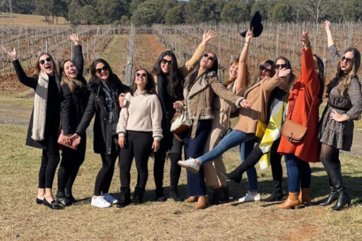 Group of joyful people posing outdoors in front of vineyard, waving and smiling.