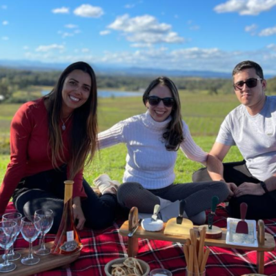 Three people sitting on a picnic blanket with wine and snacks outdoors.