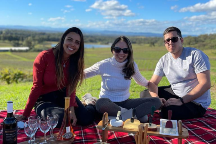 Three people sitting on a picnic blanket with wine and snacks outdoors.