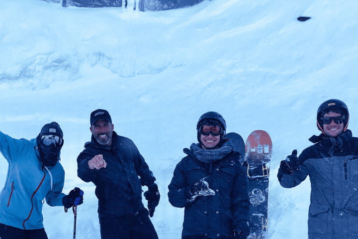 Four people in winter clothing posing with skis and a snowboard on a snowy slope.
