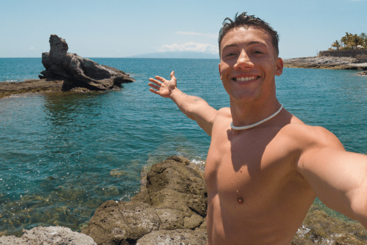 Man taking a selfie by the ocean with rocks and clear sky.