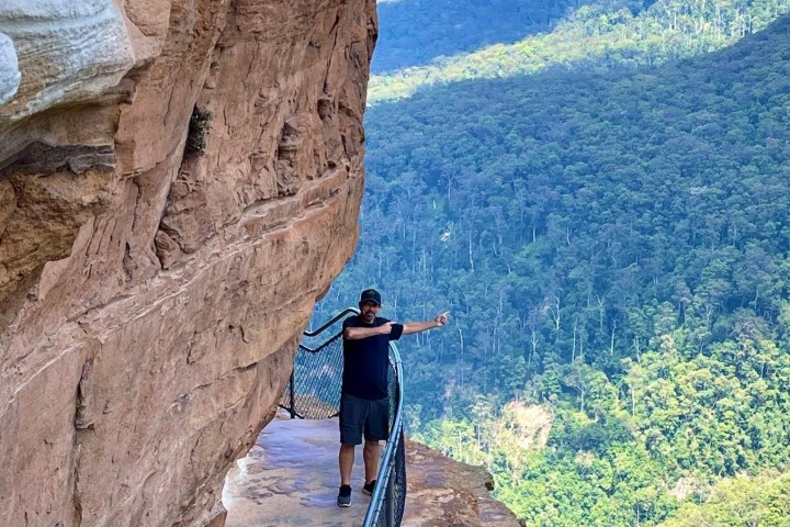Person standing on narrow cliff path overlooking green valley.