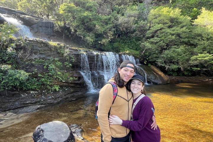 Couple hugs near a small waterfall and lush greenery.