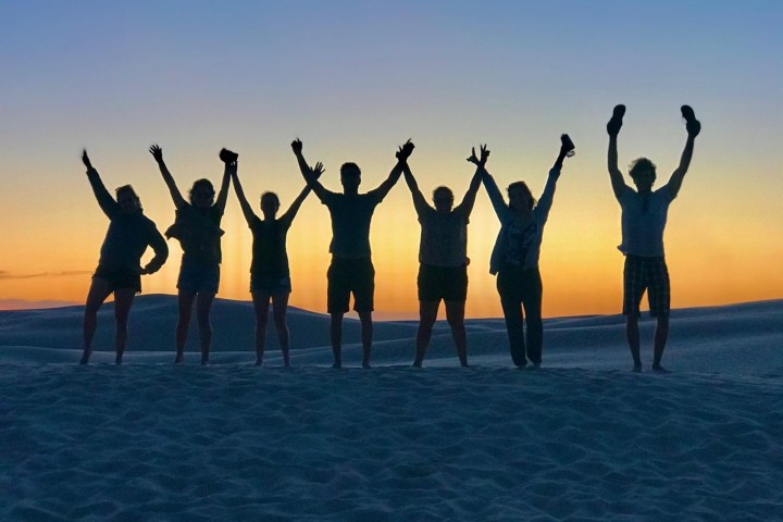 Silhouettes of seven people with raised arms at sunset on a sandy dune.