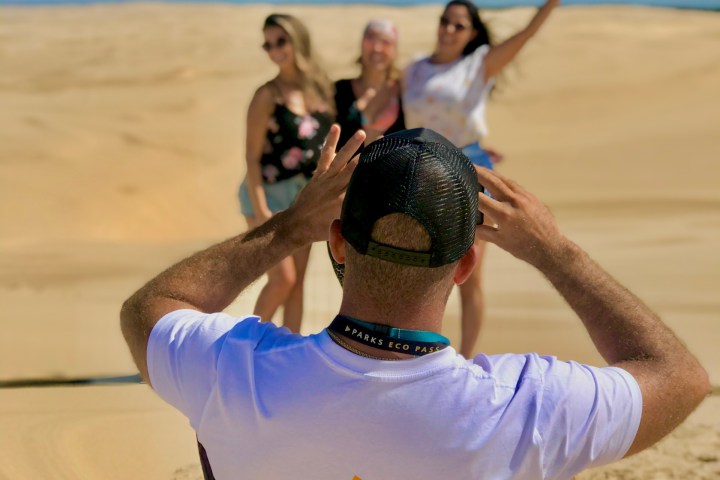 Person photographing three women on sand dunes under blue sky.