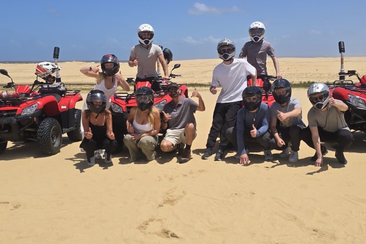 Group posing with ATVs on sandy terrain under clear blue sky.