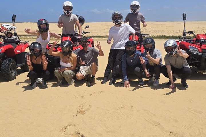 Group posing with ATVs on sand, clear blue sky in background.